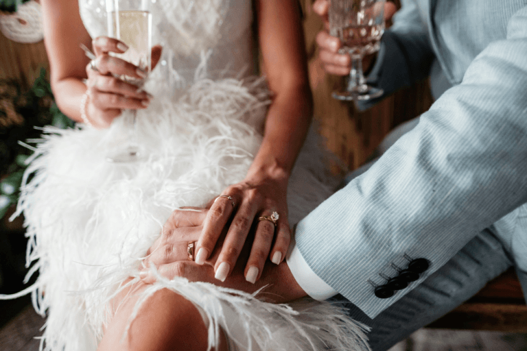 Zbliżenie na splecione dłonie pary młodej w ślubnych strojach (kobieta w sukni z pierza), trzymających kieliszki. Na dłoniach widać złote obrączki.