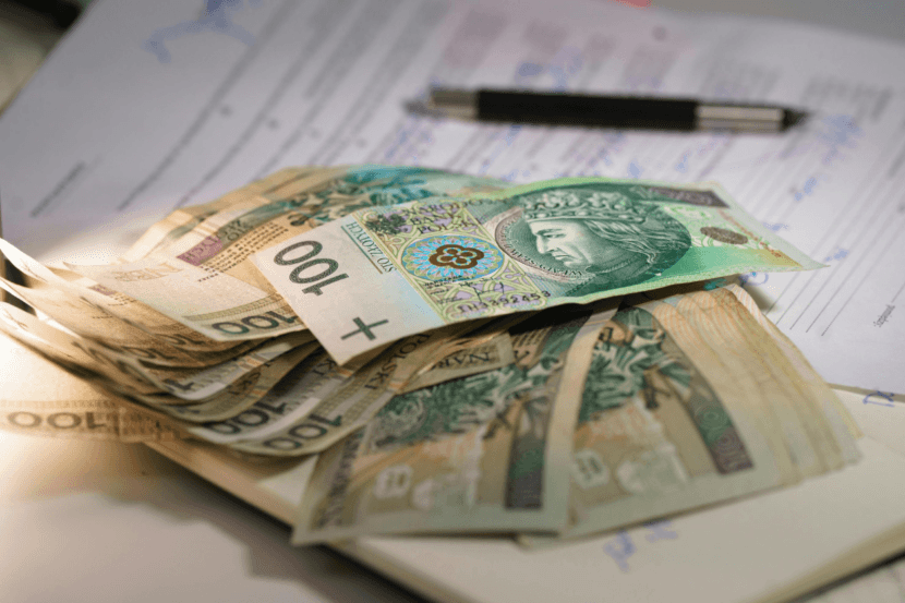 Stos polskich banknotów o nominale 100 złotych rozsypany na notatniku i dokumencie, z długopisem w tle. Wyróżnia się banknot z napisem "+100".