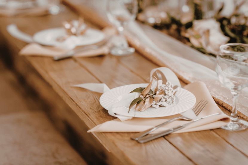 Rustykalne nakrycie weselne na drewnianym stole: biały talerz z drobną kompozycją ze złotego wieńca, gipsówki i białej wstążki.