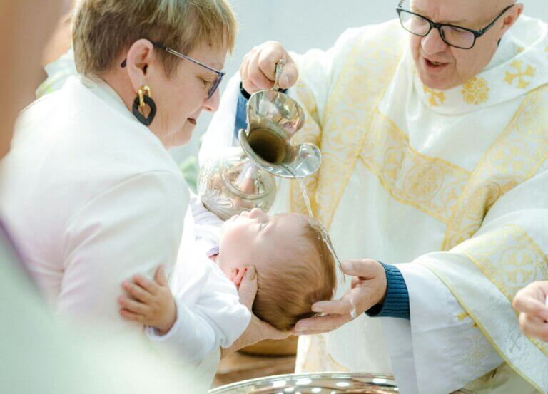 Ksiądz wlewa wodę chrzcielną na główkę niemowlęcia trzymanego przez matkę.